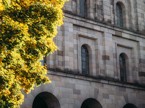 Teilansicht der Kongresshalle Nürnberg mit steinernem Fassadenteil und Rundbogenarkaden, links ein Baum mit herbstlich gefärbtem Laub.