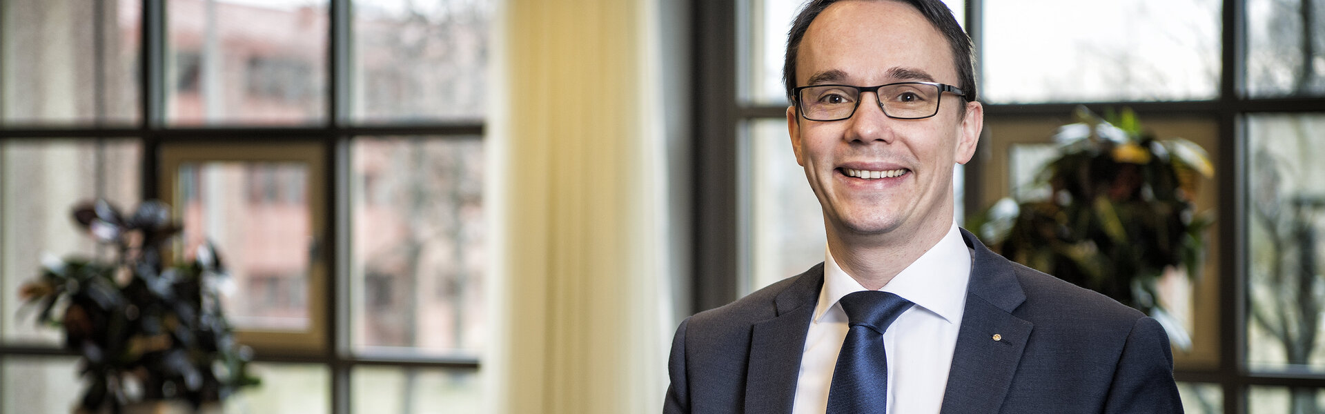 Porträtaufnahme von Jörg Schlag im Anzug mit Krawatte, der lächelnd vor einer großen Fensterfront in einem hellen Konferenz- oder Veranstaltungsraum steht.