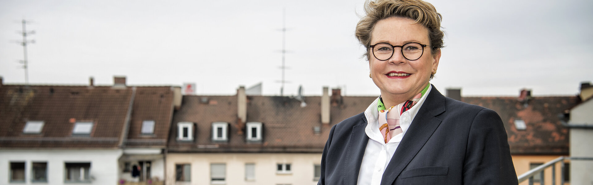 Porträtaufnahme von Sabine Unckell im dunklen Blazer vor einer städtischen Kulisse mit Wohnhäusern; sie steht lächelnd an einem Metallgeländer im Freien.