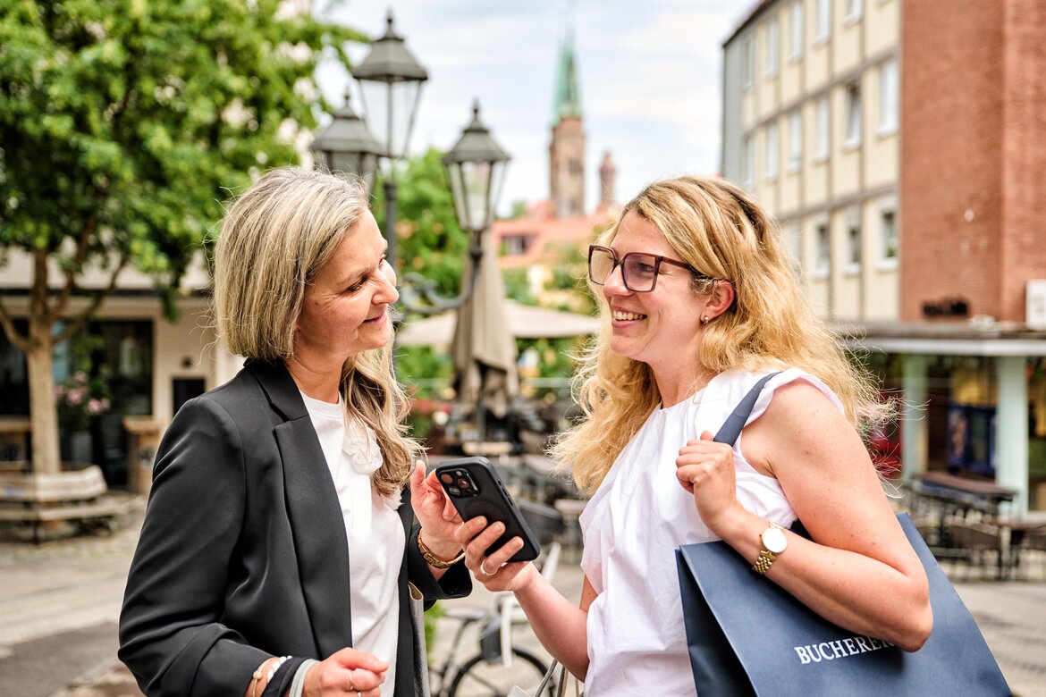 Zwei Frauen unterhalten sich lachend auf einem Platz in Nürnberg, eine hält ein Smartphone, die andere trägt eine Tasche