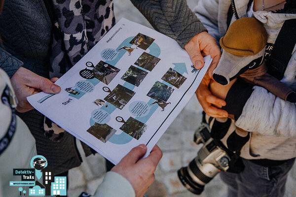 Zwei Personen, eine hält eine Detektiv-Trail-Karte mit Fotos und Texten, die andere trägt eine Kamera um den Hals, beide stehen auf gepflastertem Boden.