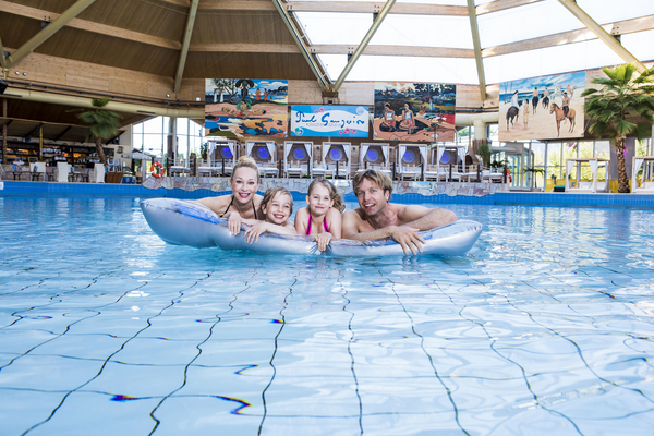 Eine vierköpfige Familie lehnt lachend auf einer silbernen Luftmatratze im großen Innenbecken des Erlebnisbads. Das Schwimmbad ist mit Palmen, Südsee-Gemälden und einer markanten Holz-Glaskuppel-Konstruktion gestaltet.