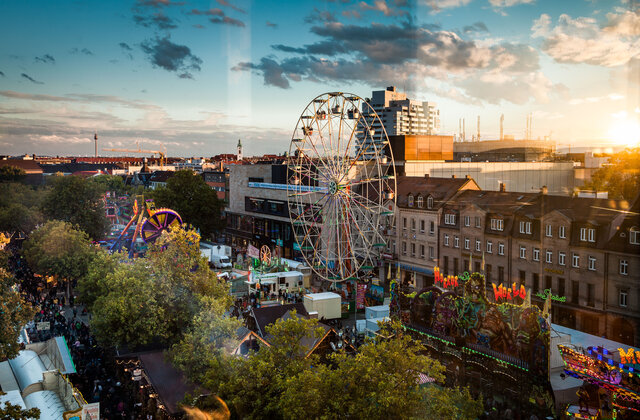 Blick von oben auf die Michaelis-Kirchweih in Fürth bei Sonnenuntergang. Ein großes Riesenrad steht im Zentrum des belebten Festplatzes, umgeben von verschiedenen Fahrgeschäften und Buden.