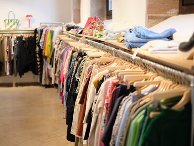 Laden mit mehreren Kleiderstangen, auf denen bunte Kleidung auf Holzkleiderbügeln hängt, und Regalen mit gefalteter Kleidung und Schuhen