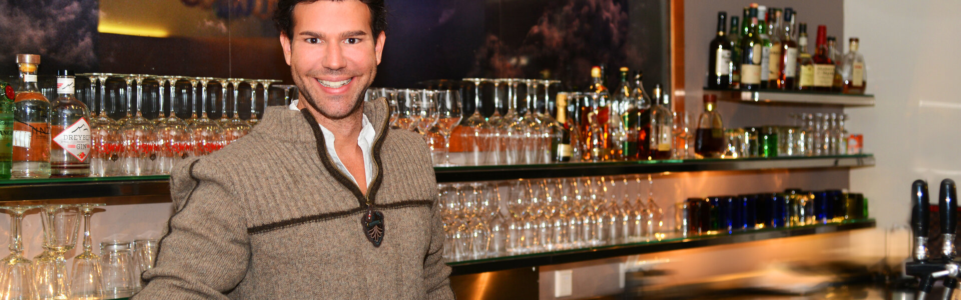 Christian Wagner mit dunklen, leicht lockigen Haaren und einem herzlichen Lächeln steht hinter einer modern beleuchteten Bar. Er trägt einen beigefarbenen Strickpullover mit Reißverschluss und einen dekorativen Anhänger am Kragen.