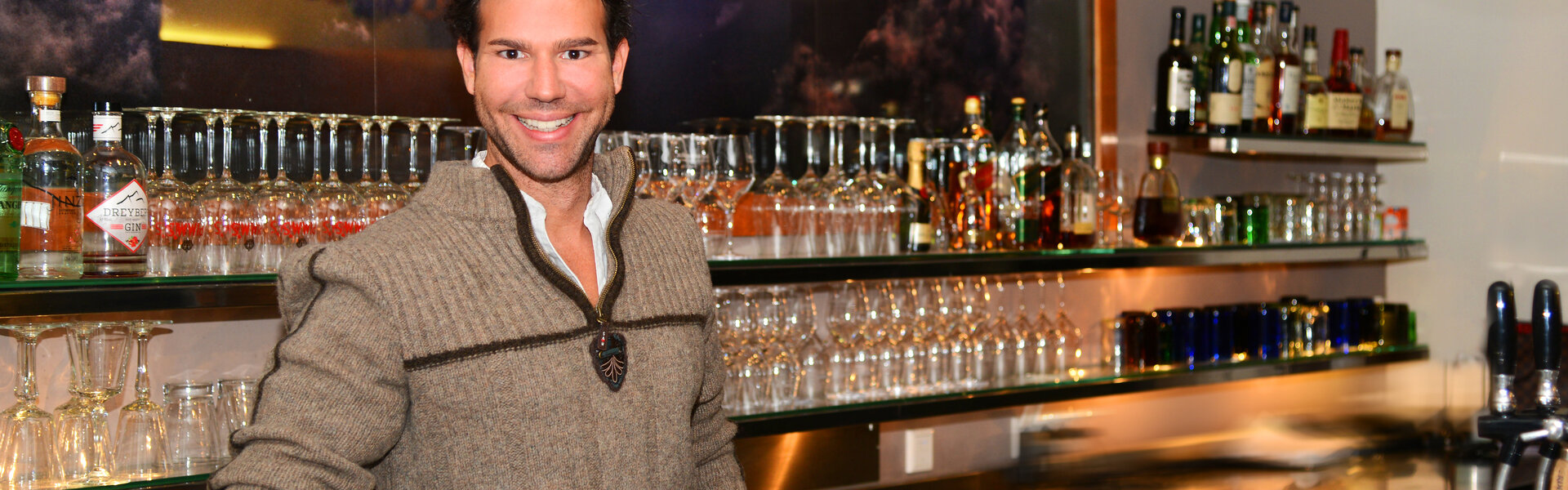 Christian Wagner mit dunklen, leicht lockigen Haaren und einem herzlichen Lächeln steht hinter einer modern beleuchteten Bar. Er trägt einen beigefarbenen Strickpullover mit Reißverschluss und einen dekorativen Anhänger am Kragen.