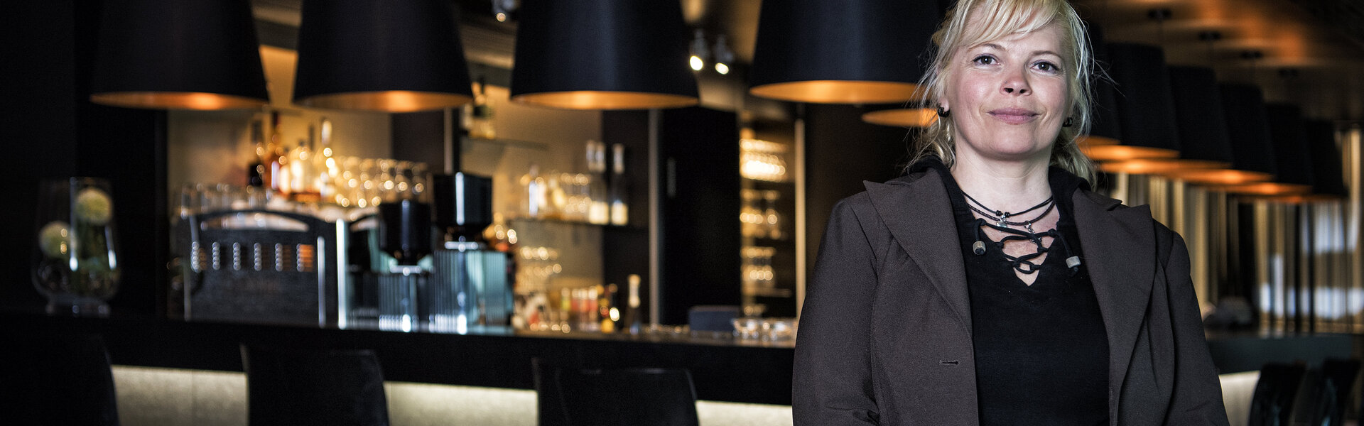 Porträtaufnahme von Monika Scheffler im braunen Mantel vor einer stilvoll beleuchteten, modernen Bar mit hohen Hockern und großen schwarzen Hängelampen.