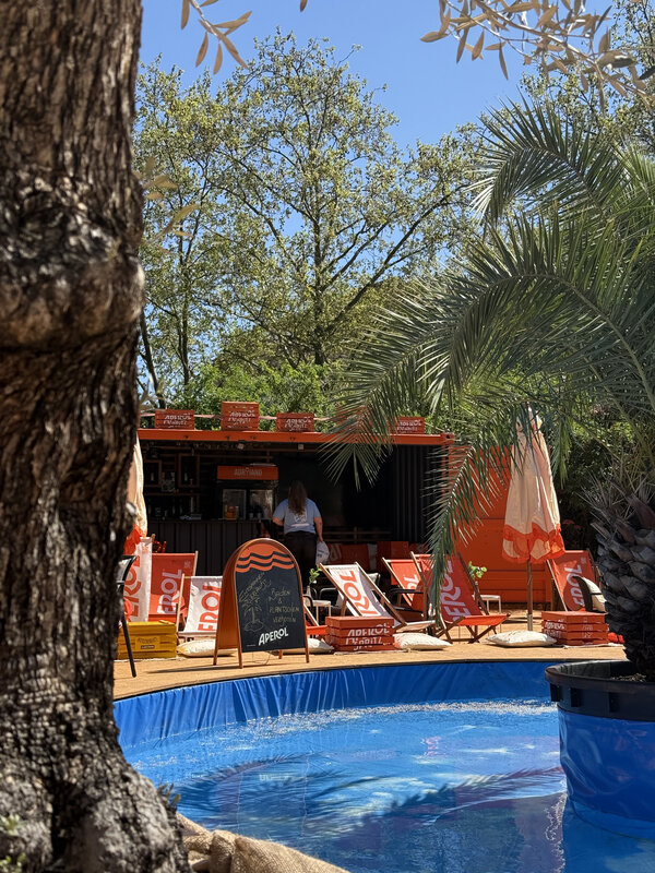 Blick auf einen kleinen Pool mit blauem Rand vor einer Bar mit orangen Sonnenschirmen und Liegestühlen, umgeben von Bäumen und Palmen auf der Sommerinsel Nürnberg