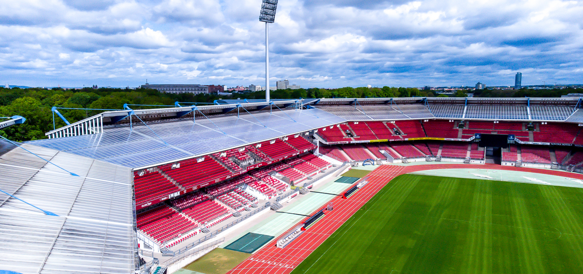 Luftaufnahme des Nürnberger Max-Morlock-Stadions: Der Blick schwebt über die markanten, eckigen Tribünen mit ihren roten Sitzreihen und dem grünen Spielfeld. Über dem weiten Stadionrund ragt ein hoher Flutlichtmast in den bewölkten Himmel.