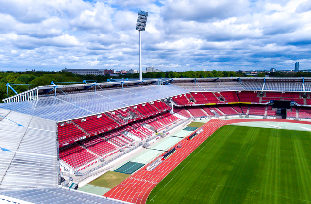 Luftaufnahme des Nürnberger Max-Morlock-Stadions: Der Blick schwebt über die markanten, eckigen Tribünen mit ihren roten Sitzreihen und dem grünen Spielfeld. Über dem weiten Stadionrund ragt ein hoher Flutlichtmast in den bewölkten Himmel.