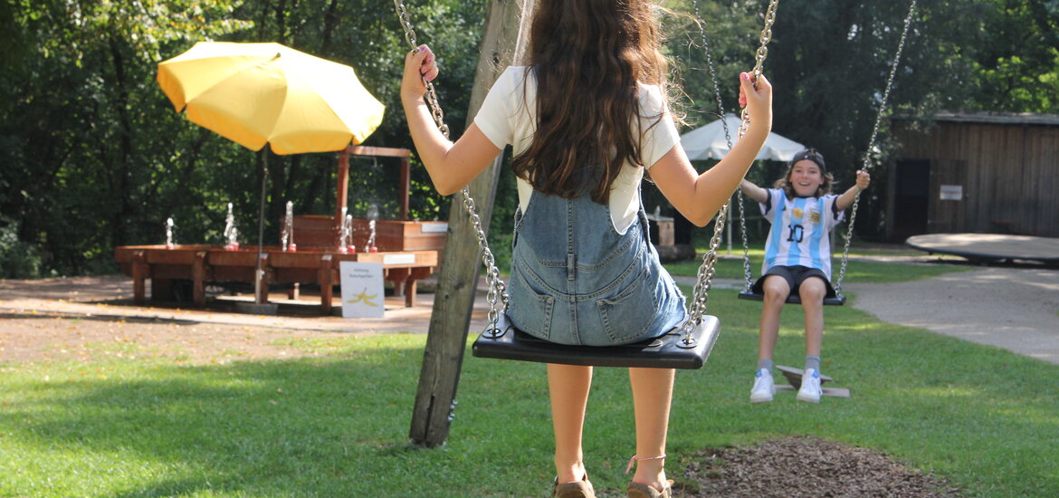 Zwei Kinder schaukeln auf einer Doppelschaukel im Erfahrungsfeld Nürnberg, im Hintergrund ein gelber Sonnenschirm und eine Sitzgruppe auf einer Wiese