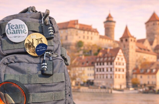 Grauer Rucksack von Team Game mit mehreren Vorhängeschlössern, zwei runden Ansteckbuttons und einer Lupe vor einer Kulisse mit dem Fluss Pegnitz, historischen Gebäuden und der Kaiserburg..