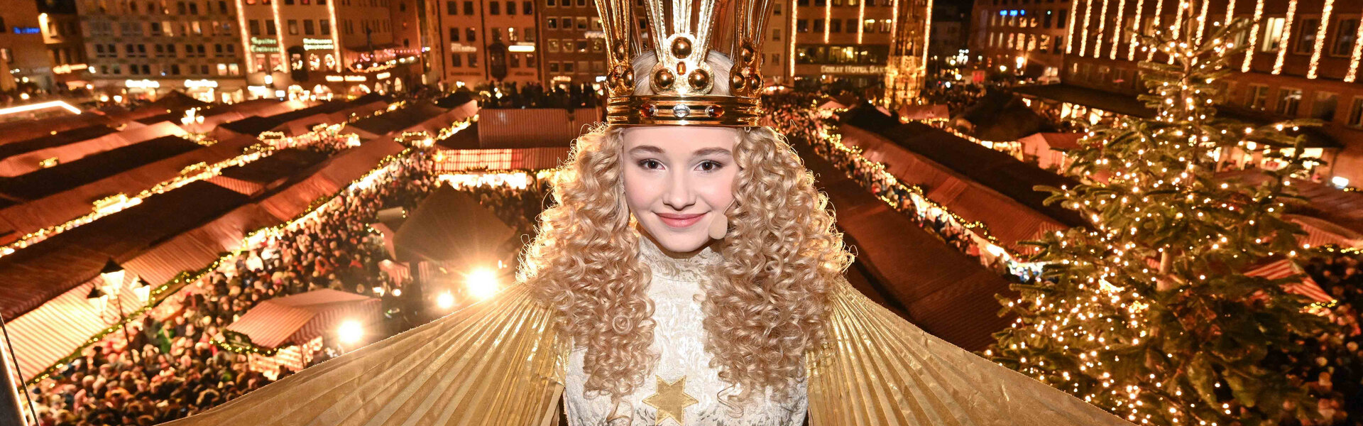 Das Nürnberger Christkind mit goldener Krone und lockigem Haar steht vor dem beleuchteten Christkindlesmarkt mit festlich geschmückten Ständen und Weihnachtsbäumen