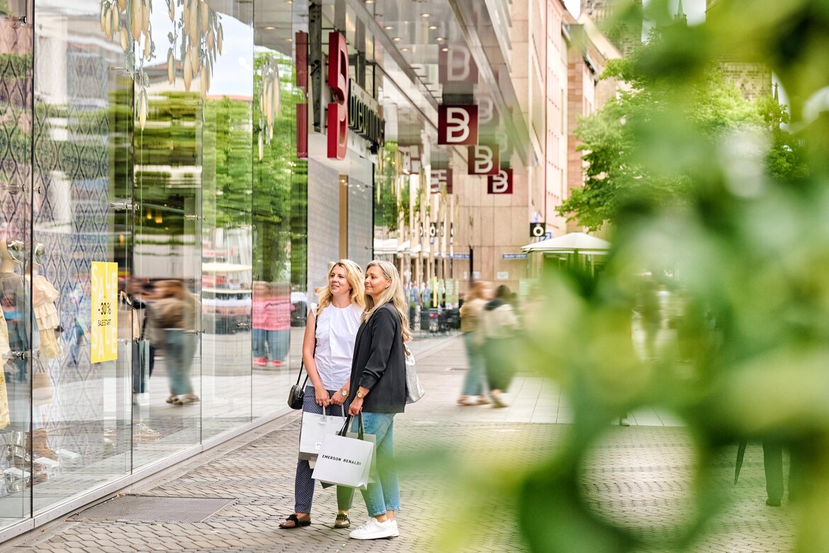 Zwei Frauen mit Einkaufstüten stehen vor einem Schaufenster in der Karolinenstraße Nürnberg, umgeben von modernen Ladenfassaden und Passanten.