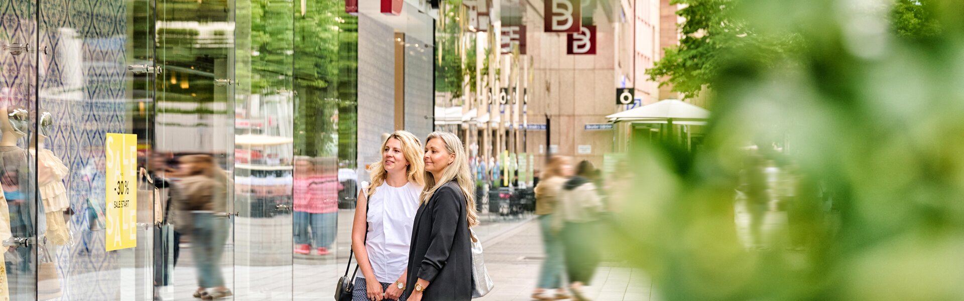 Zwei Frauen mit Einkaufstüten stehen vor einem Schaufenster in der Karolinenstraße Nürnberg, umgeben von modernen Ladenfassaden und Passanten.