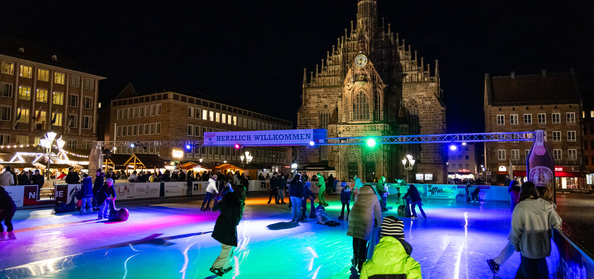 Eislaufende Menschen auf der beleuchteten Eisfläche vor der beleuchteten Frauenkirche am Nürnberger Hauptmarkt bei Nacht, darüber ein Banner mit der Aufschrift 'HERZLICH WILLKOMMEN'.