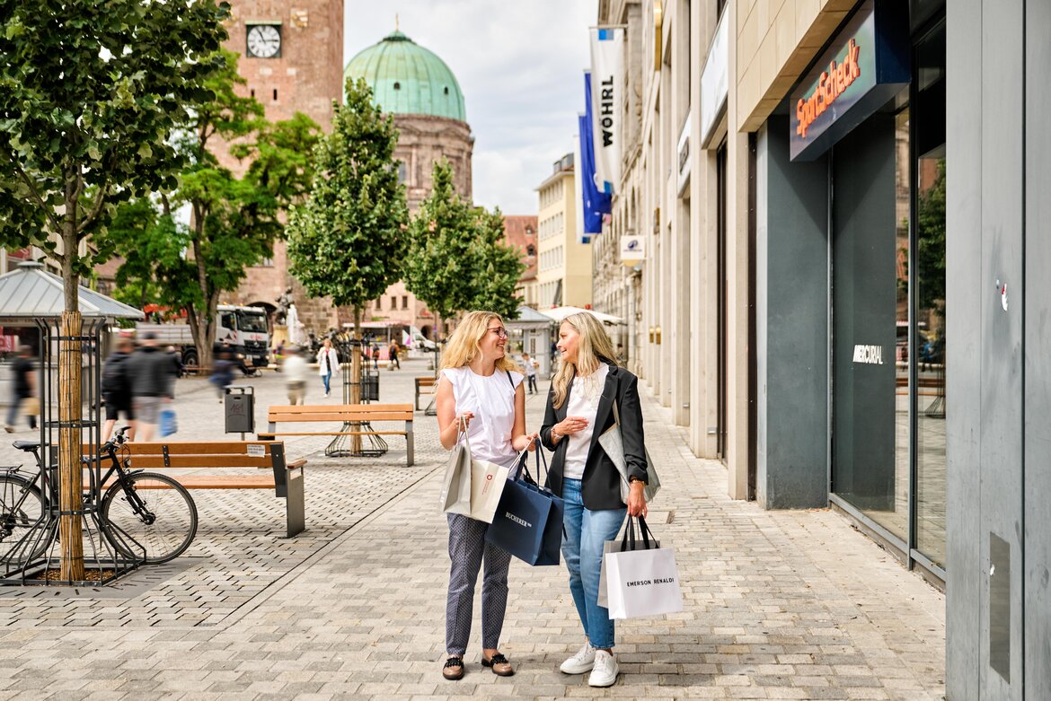 Zwei Frauen mit Einkaufstüten unterhalten sich auf dem gepflasterten Ludwigsplatz in Nürnberg vor Geschäften und dem Weißen Turm und der Elisabethkircheim Hintergrund