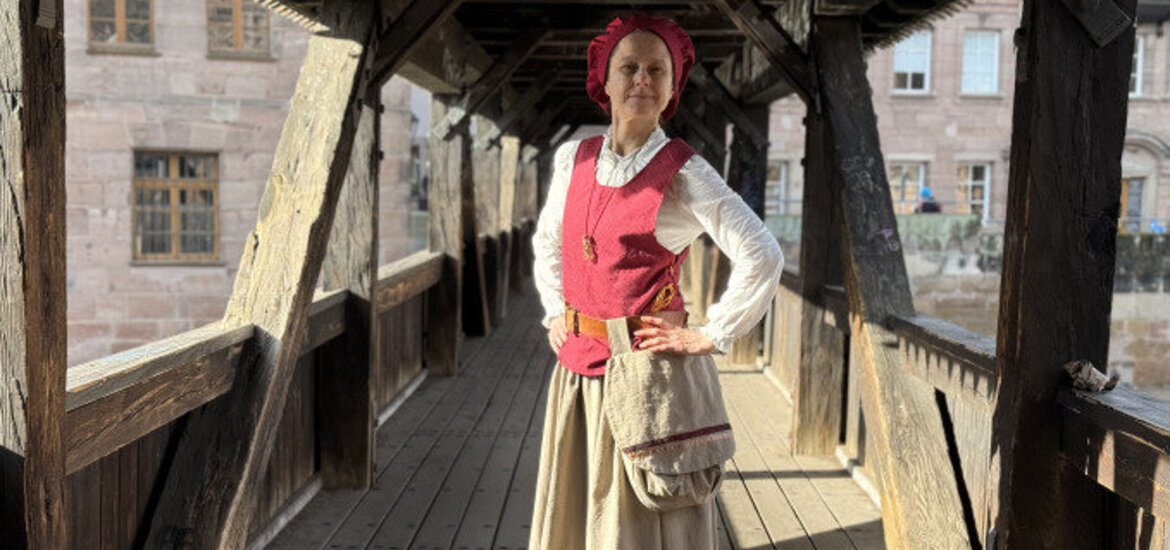 Eine als Henkersfrau Maria Schmidt verkleidete Dame steht im historischen Gewand mitten auf dem Henkersteg und lächelt in die Kamera.