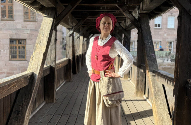 Eine als Henkersfrau Maria Schmidt verkleidete Dame steht im historischen Gewand mitten auf dem Henkersteg und lächelt in die Kamera.