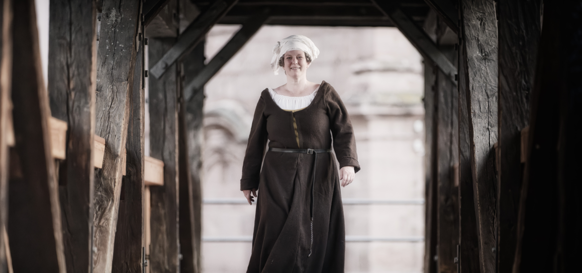 Frau in mittelalterlicher Kleidung mit weißer Haube geht auf einem hölzernen überdachten Steg (Henkersteg)