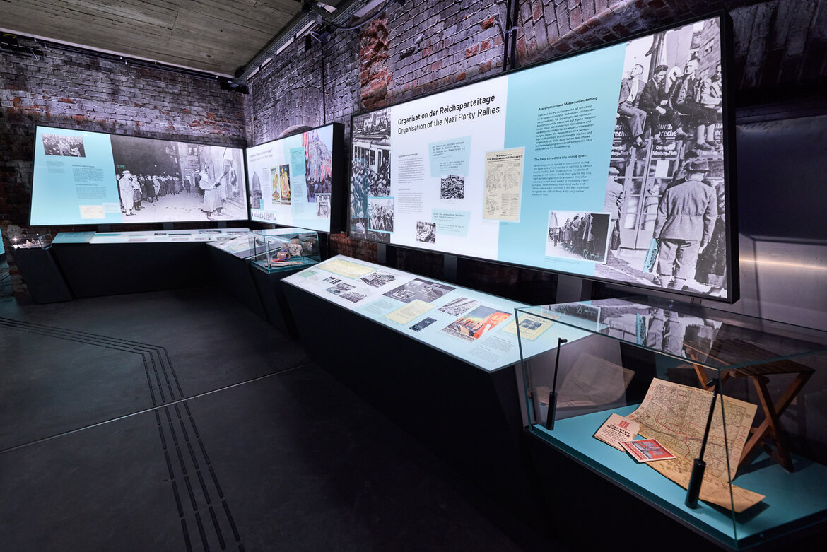 Ausstellungsraum der neuen Dauerausstellung im Dokuzentrum Nürnberg mit beleuchteten Informationstafeln, historischen Fotos und Dokumenten in Vitrinen vor einer rohen Backsteinwand