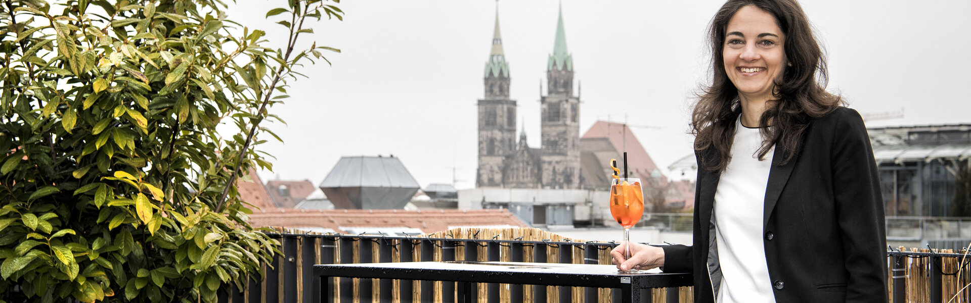 Frau steht auf einer Dachterrasse mit Getränk in der Hand, umgeben von modernen Möbeln, im Hintergrund historische Stadtsilhouette mit Doppelturm‑Kirche unter grauem Himmel.