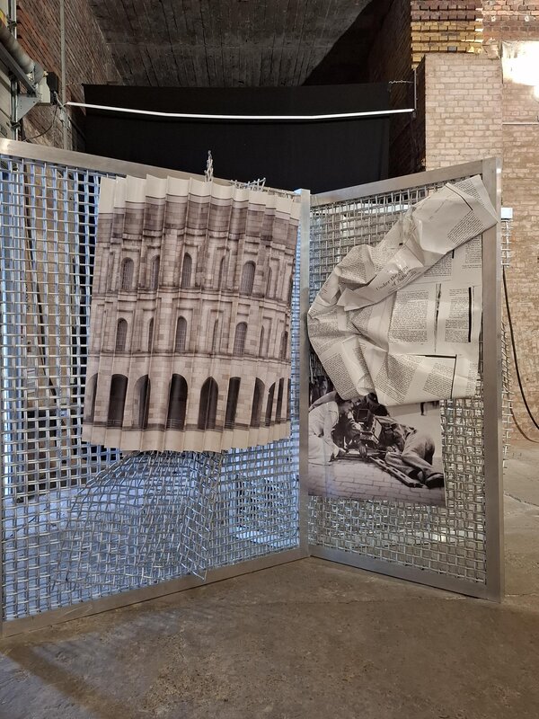 Metallgitter mit zwei daran befestigten bedruckten Papierbögen, einer mit einem architektonischen Motiv und der andere zerknüllt mit Text und einem Bild von einer Statue