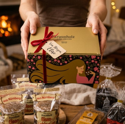 Hände halten eine goldfarbene Geschenkbox mit roter Schleife und Aufdruck einer Frau mit langen Haaren, umgeben von verpackten Lebkuchenprodukten auf Holztisch vor Kaminfeuer