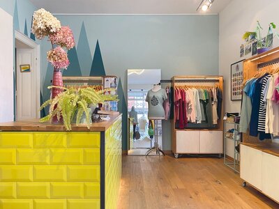 Innenraum eines kleinen Bekleidungsgeschäfts mit gelber Kacheltheke, hölzernem Boden, Pflanzen auf der Theke und Kleidung an Holzständern