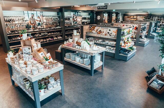 Großzügige Innenansicht eines modernen Fackelmann Outlets: Auf hellen Holztischen und in dunklen Regalsystemen wird ein breites Sortiment an Küchenhelfern, weißem Geschirr und Backzubehör präsentiert.