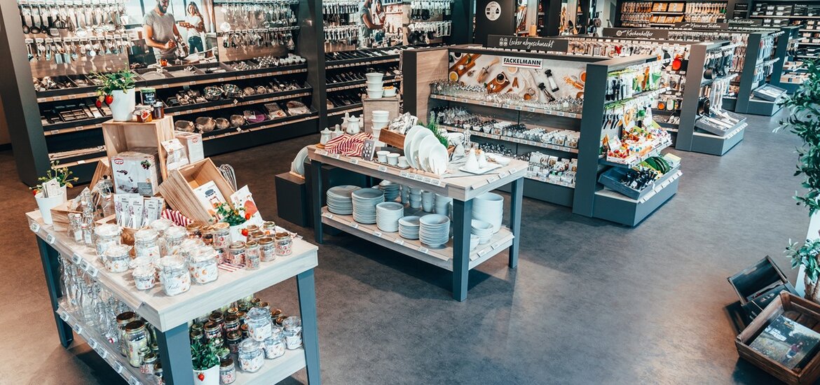 Großzügige Innenansicht eines modernen Fackelmann Outlets: Auf hellen Holztischen und in dunklen Regalsystemen wird ein breites Sortiment an Küchenhelfern, weißem Geschirr und Backzubehör präsentiert.