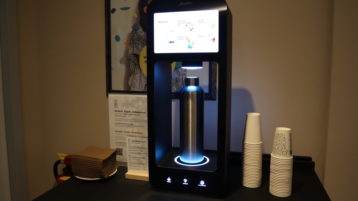 Intelligenter Wasserspender mit beleuchtetem Flaschenhalter, Touch-Bedienfeld und Display, daneben Pappbecher und Servietten auf schwarzem Tisch.