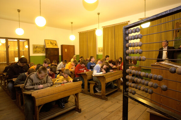 Ein historisches Klassenzimmer im Schulmuseum, in dem Kinder an alten Holzschulbänken schreiben, während im Vordergrund eine große Rechenmaschine mit Holzkugeln zu sehen ist.