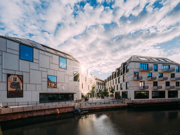Moderner Gebäudekomplex mit weißer Fassade und vielen Fenstern am Ufer eines Flusses mit Bäumen auf dem gepflasterten Innenhof und Spiegelung im Wasser der Pegnitz im Augustinerhof Nürnberg