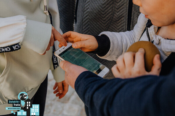 Kind und Erwachsener halten gemeinsam ein Smartphone, auf dem Text sichtbar ist, im Hintergrund eine graue Jacke und ein helles Sweatshirt mit schwarzem Schriftzug.