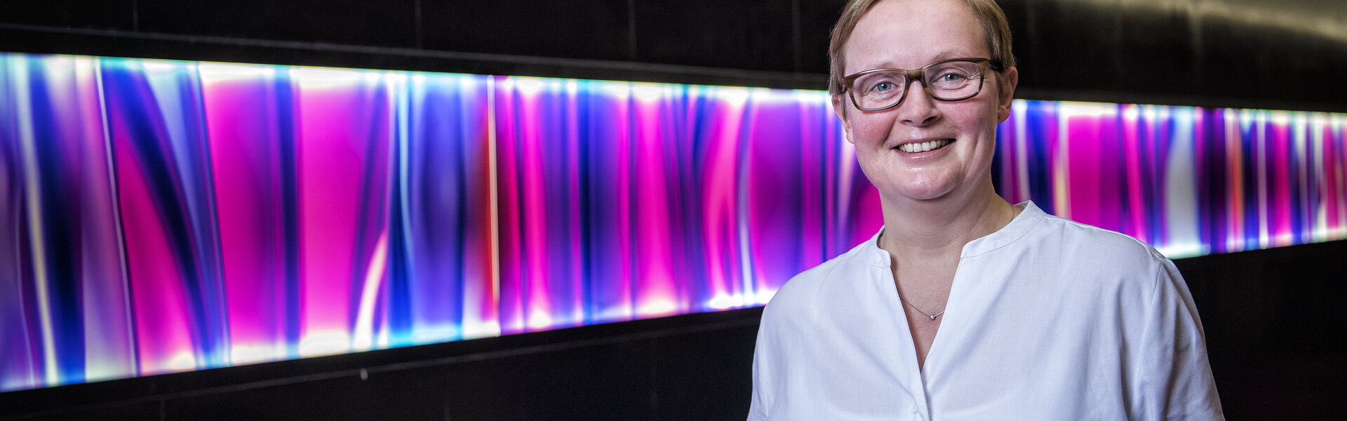 Porträtaufnahme von Karin Schuldenzucker in weißer Bluse vor einer dunklen Wand mit einem leuchtenden, violett-blauen abstrakten Lichtband.