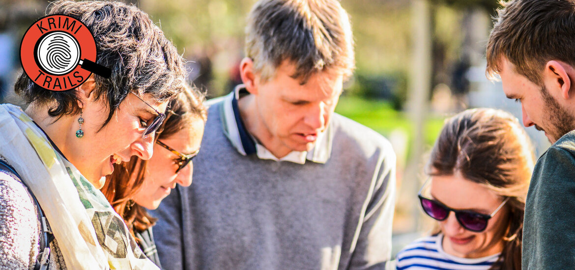 Gruppe von fünf Personen steht im Freien und betrachtet gemeinsam ein Blatt Papier und ein Smartphone, im Vordergrund rot-weiße Absperrbänder und oben links ein rundes Logo mit der Aufschrift 'KRIMI TRAILS'