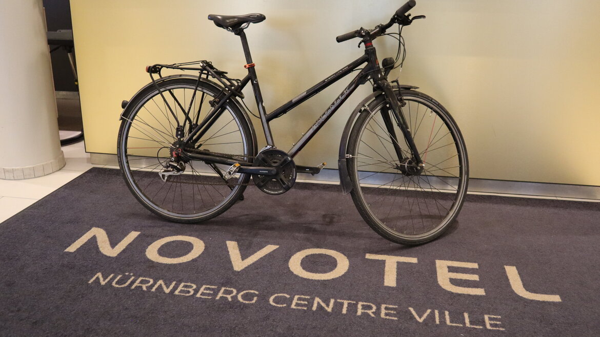 Schwarzes Fahrrad mit Gepäckträger steht auf einem Teppich mit der Aufschrift 'NOVOTEL NÜRNBERG CENTRE VILLE' vor einer hellen Rezeptionstheke.