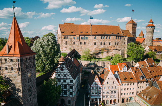 Eine Luftaufnahme der Nürnberger Kaiserburg mit ihren markanten Türmen und dem Palas über den historischen Fachwerkhäusern und roten Ziegeldächern des Burgviertels bei strahlendem Sonnenschein.
