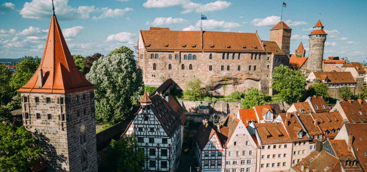 Eine Luftaufnahme der Nürnberger Kaiserburg mit ihren markanten Türmen und dem Palas über den historischen Fachwerkhäusern und roten Ziegeldächern des Burgviertels bei strahlendem Sonnenschein.