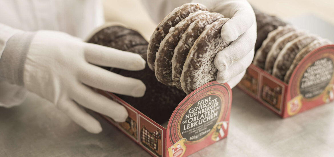 Hände in weißen Handschuhen legen frisch glasierte Nürnberger Elisen-Lebkuchen von Lebkuchen Schmidt sorgfältig in eine rote Verkaufsverpackung mit der Aufschrift „Feine gemischte Nürnberger Oblaten-Lebkuchen“.