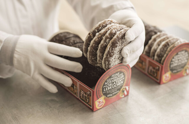 Hände in weißen Handschuhen legen frisch glasierte Nürnberger Elisen-Lebkuchen von Lebkuchen Schmidt sorgfältig in eine rote Verkaufsverpackung mit der Aufschrift „Feine gemischte Nürnberger Oblaten-Lebkuchen“.
