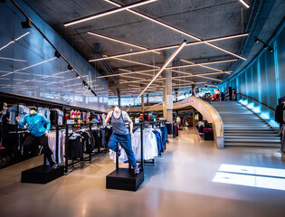 Moderner, weitläufiger Verkaufsraum des Adidas Outlets mit dynamisch angeordneten Lichtröhren an der Betondecke, Sportbekleidung auf Kleiderständer und einer großen Treppe im Hintergrund.