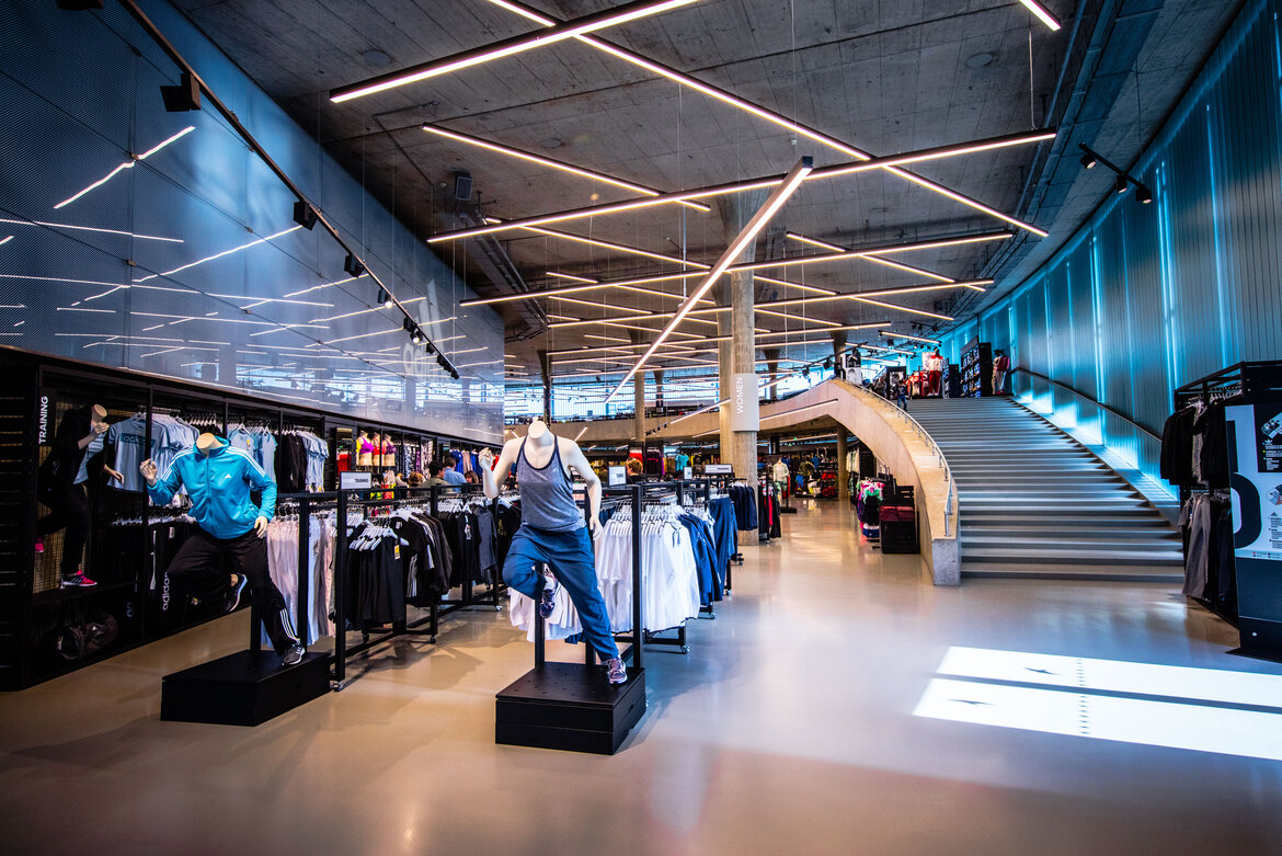A modern, spacious sales floor at the Adidas Outlet, featuring dynamically arranged fluorescent lights on the concrete ceiling, sportswear on racks, and a large staircase in the background.
