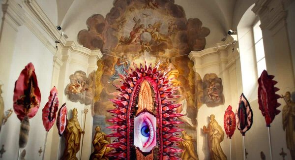 Kirchenschiff mit barocker Deckenmalerei und moderner Installation aus roten, blütenartigen Skulpturen mit einem großen Auge in der Mitte