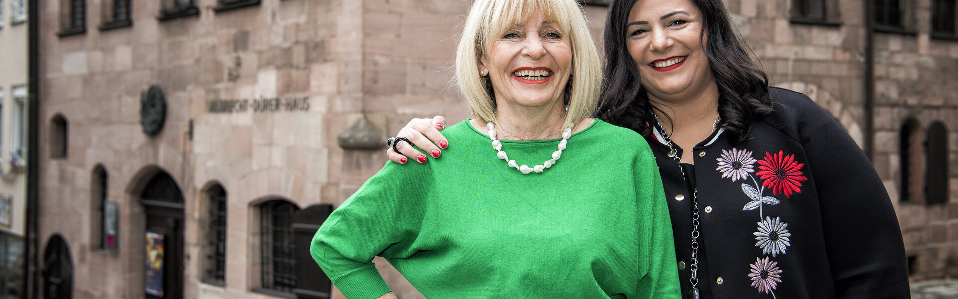 Hermine Schenkel und Yesim Dinsel stehen lächelnd Arm in Arm auf dem Kopfsteinpflaster vor dem historischen Albrecht-Dürer-Haus in Nürnberg.