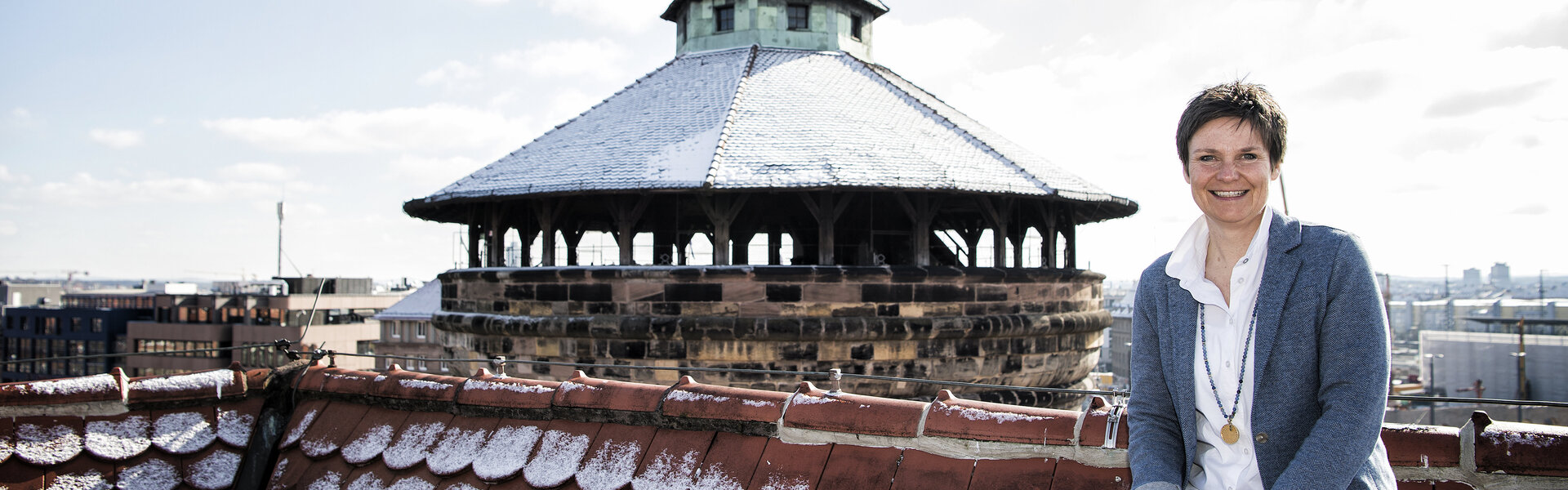 Sabine Powels sitzt lächelnd auf einem schneebedeckten Ziegeldach; im Hintergrund ist ein historischer Turm mit schneebedeckter Haube zu sehen.