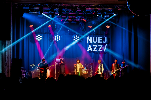 Eine mehrköpfige Jazz-Band tritt auf einer blau und rot beleuchteten Bühne auf, während helle Scheinwerferkegel den Raum kreuzen. Im Hintergrund ist der Schriftzug „NUEJAZZ“ in großen weißen Buchstaben zu sehen.