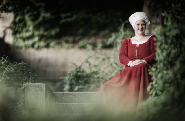 Eine lächelnde Frau in rotem mittelalterlichem Kleid und weißer Haube sitzt auf einer Steinmauer vor grünem Laub