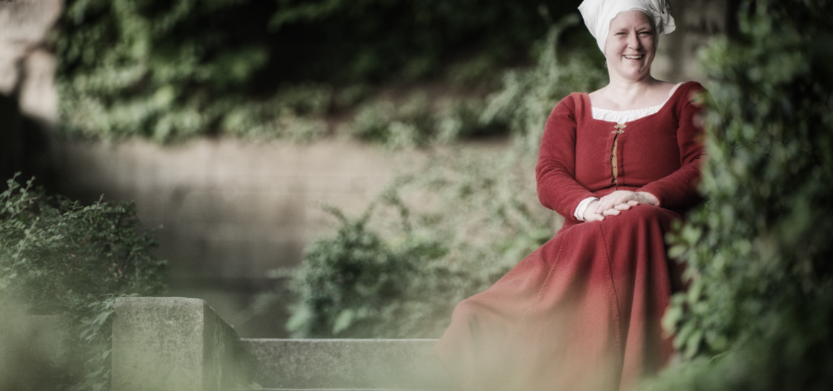 Eine lächelnde Frau in rotem mittelalterlichem Kleid und weißer Haube sitzt auf einer Steinmauer vor grünem Laub